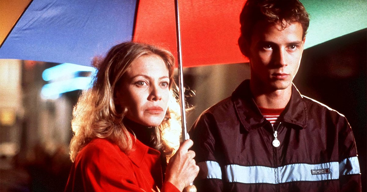 "Scène du film 'Tout sur ma mère' de Pedro Almodóvar, montrant une femme en manteau rouge et un jeune homme sous un parapluie multicolore, dans une ambiance de nuit."